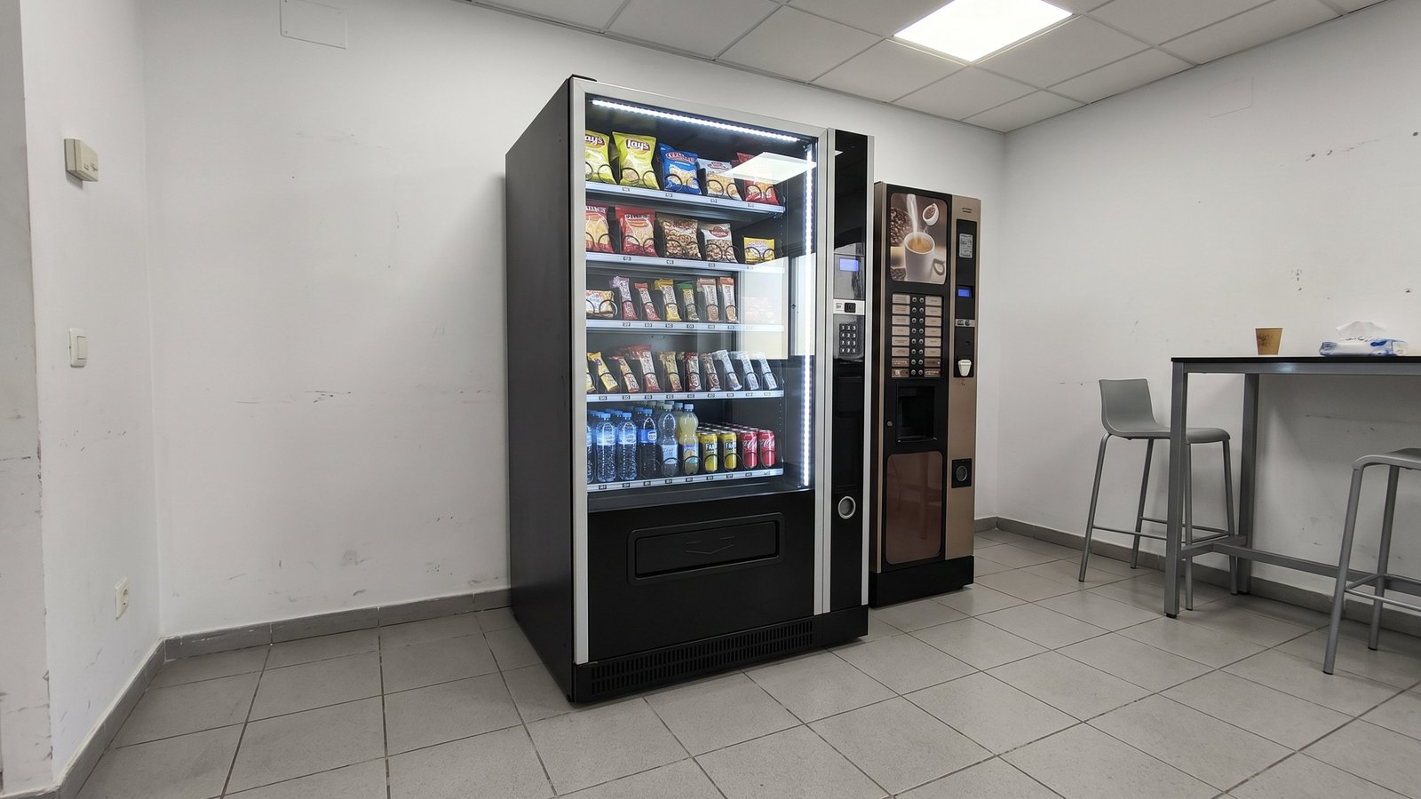 Máquina de vending mixta para empresas con snacks y bebidas frías junto a una máquina de café en la sala de descanso de una oficina